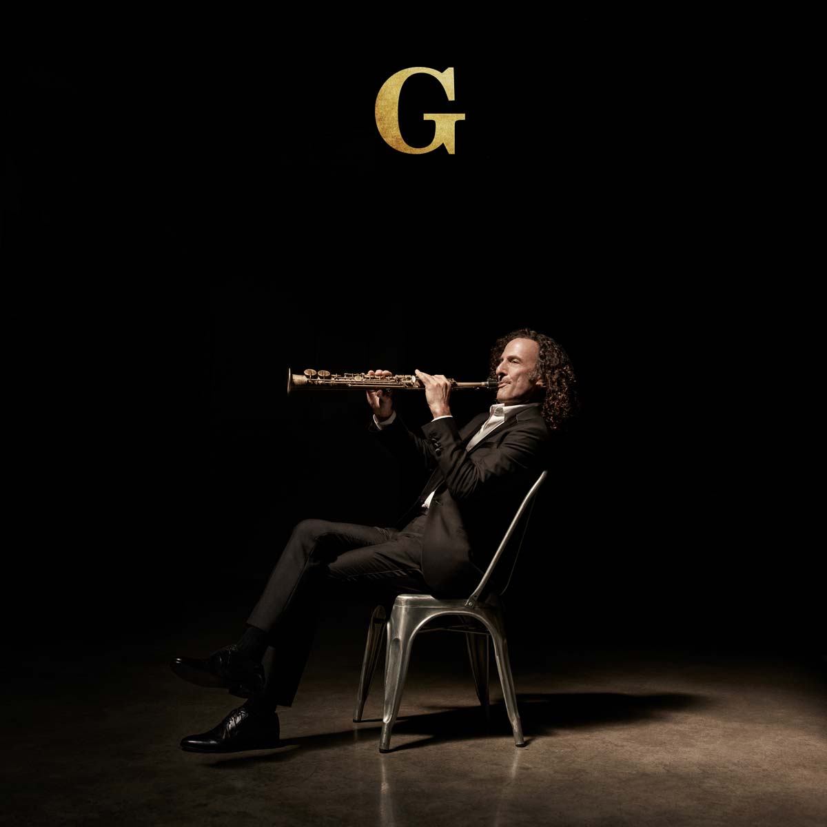 Man playing a flute on a chair with a dark background and a large 'G' above