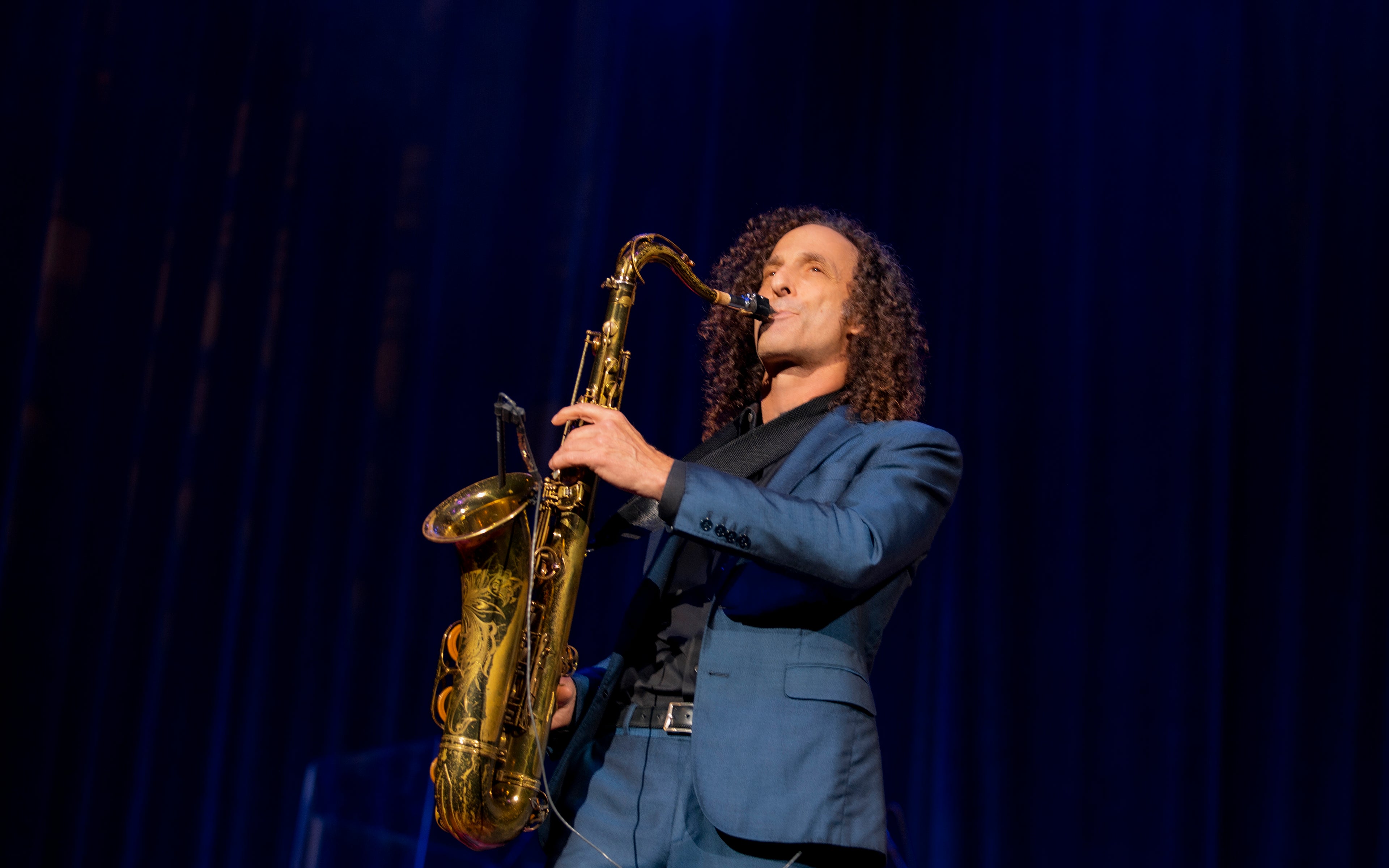 Kenny G in front of a crimson blue curtain playing his brass sax